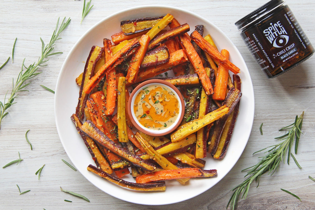 Rainbow carrot fries piled on a white plate with a tahini chili crisp dip, rosemary and a jar of Spice Witch Black Garlic Chili Crisp