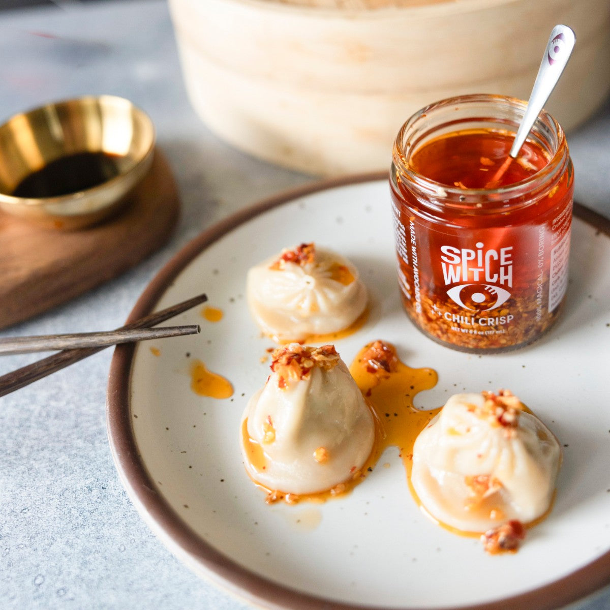 Spice Witch Original Chili Crisp jar made with avocado oil next to soup dumplings drizzled with chili crisp oil