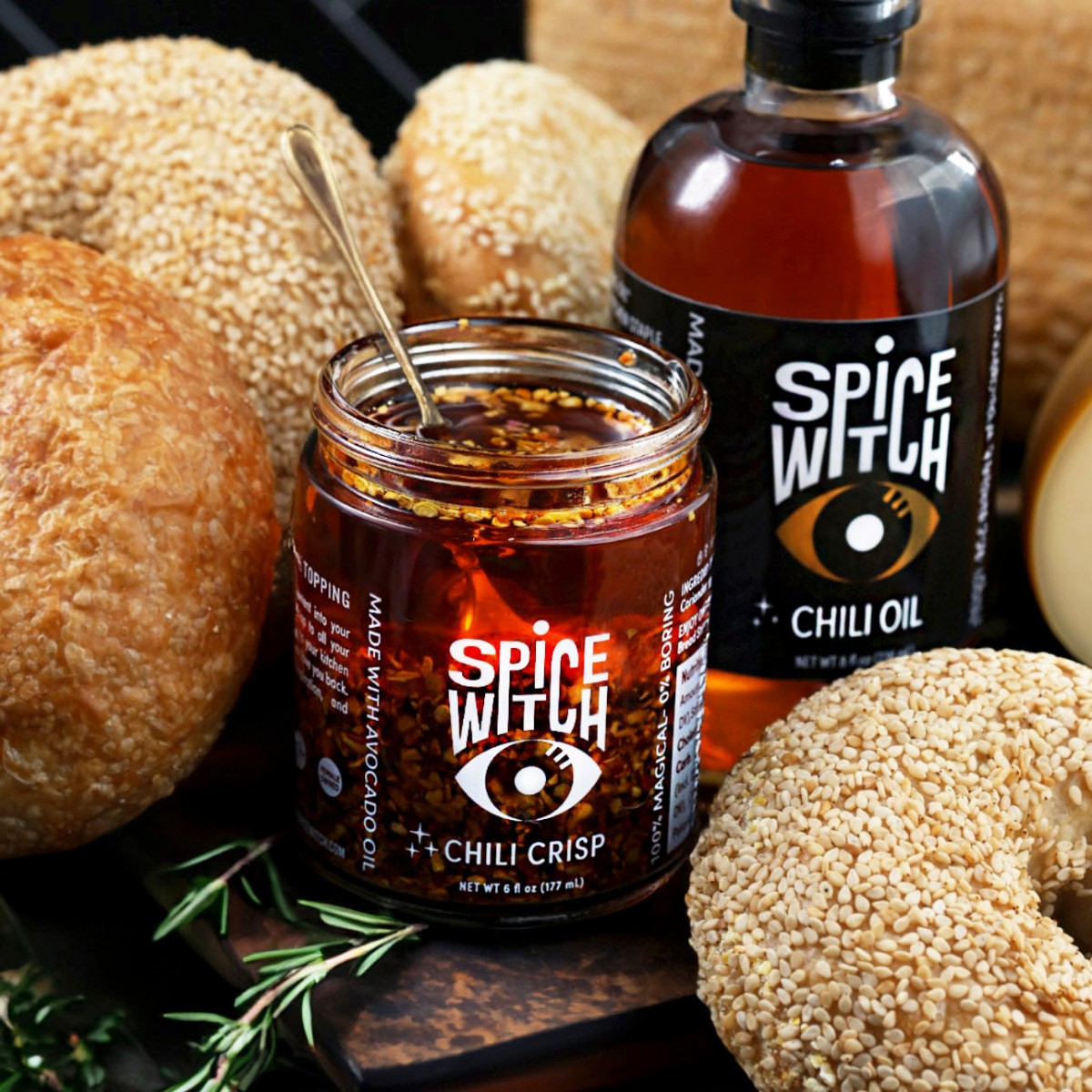 Spice Witch Original Chili Crisp jar next to a bottle of Spice Witch Chili Oil, surrounded by sesame bagels