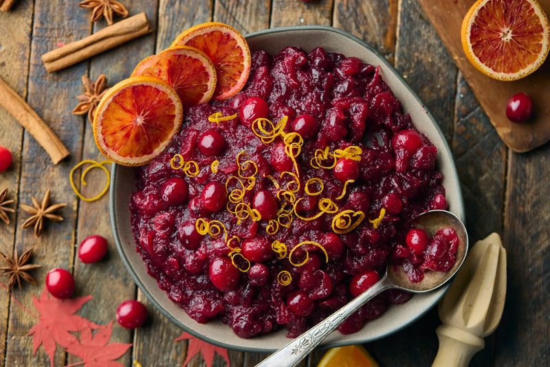 Homemade cranberry sauce in a bowl topped with orange zest and blood orange slices, with cinnamon sticks and star anise on a wooden table