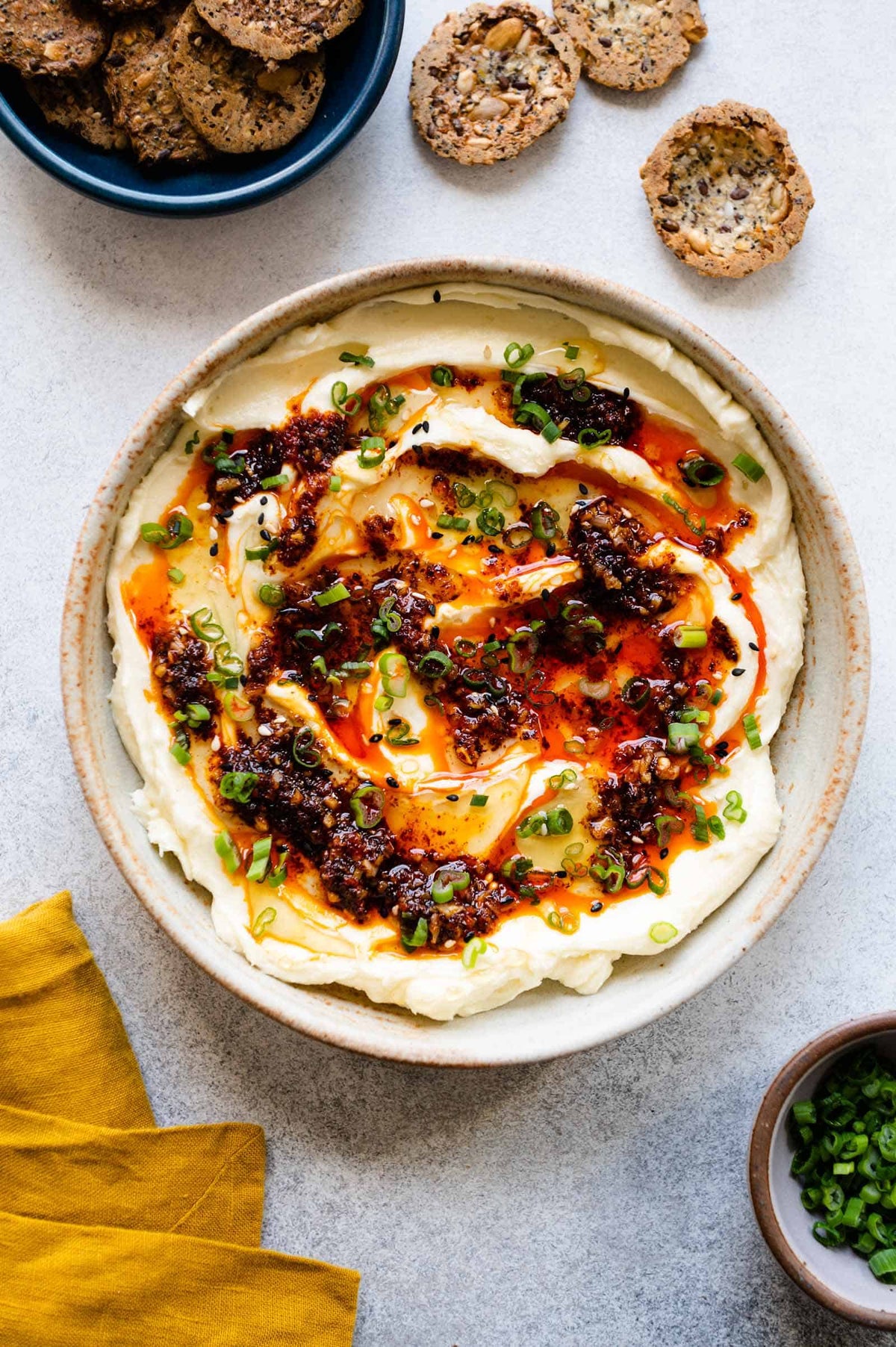 Whipped brie in a shallow bowl topped with chili crisp, hot honey, scallions and sesame seeds, with seeded crackers alongside