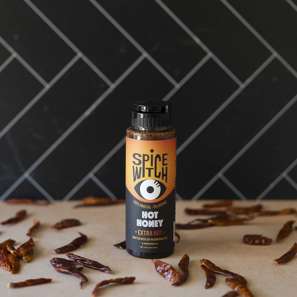 Spice Witch Extra Hot Honey bottle surrounded by dried chili peppers against black herringbone tile