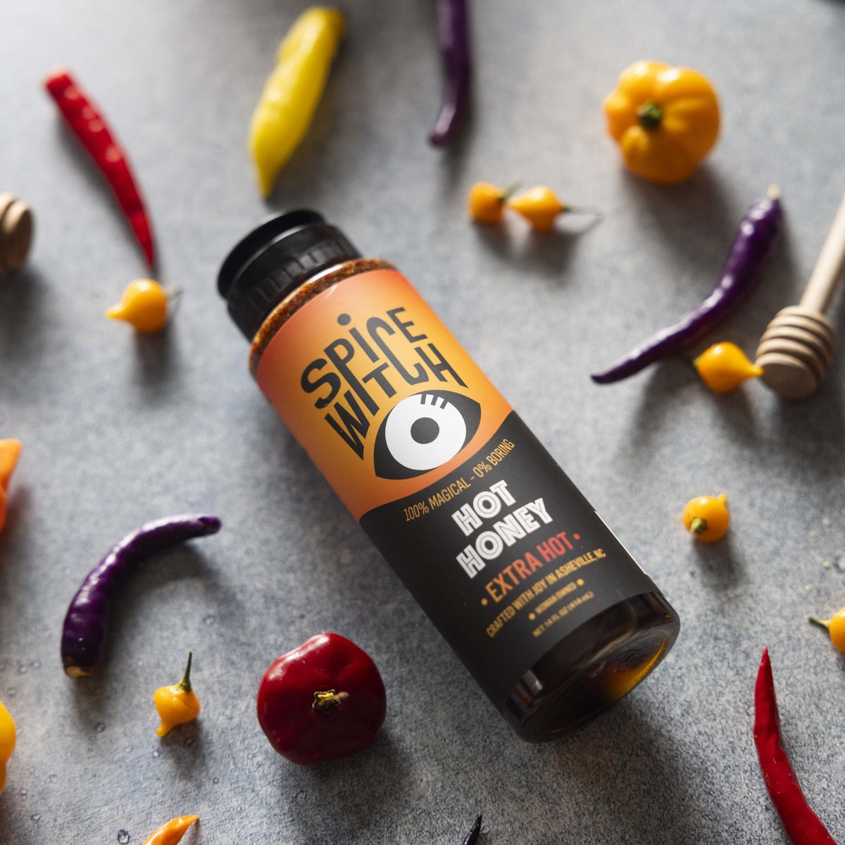 Spice Witch Extra Hot Honey 14 fl oz squeeze bottle surrounded by fresh red, yellow and purple chili peppers